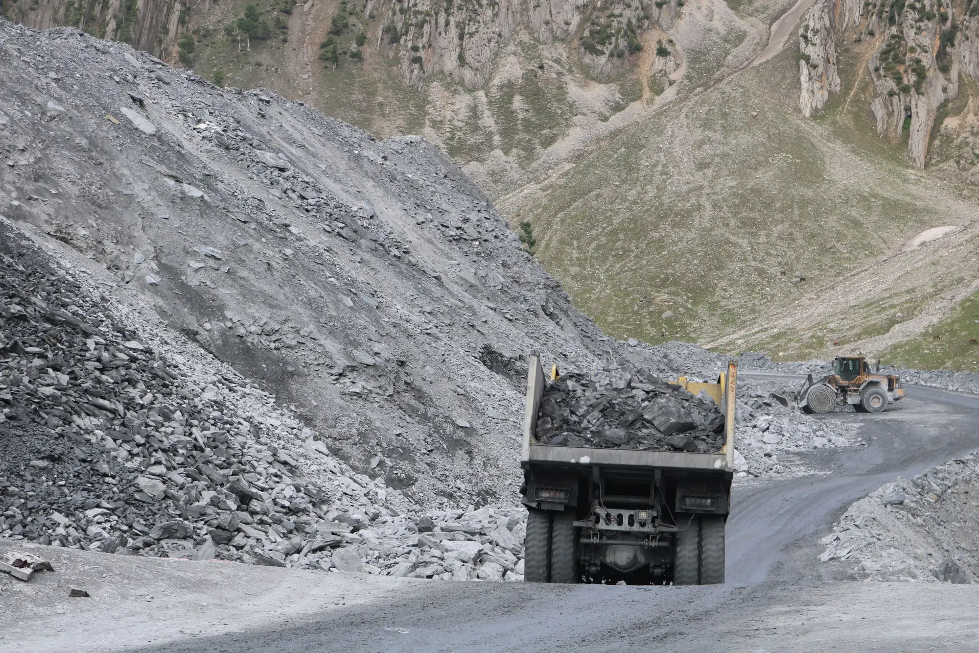 A loaded haul truck driving away up a gravel haul road between waste rock piles, a loader working behind it